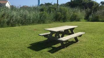 Ein Holztisch für ein Picknick in einer grünen Umgebung, mit blauem Himmel und etwas Vegetation im Hintergrund.