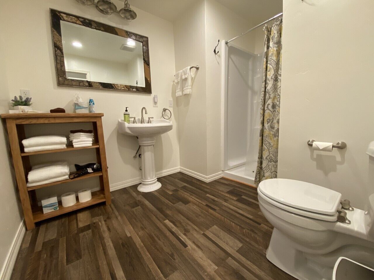 A bathroom with a wooden shelf, pedestal sink, toilet, and shower curtain.
