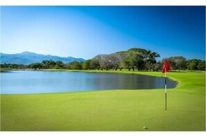 Um campo de golfe com um lago, uma bandeira vermelha e uma montanha ao fundo.