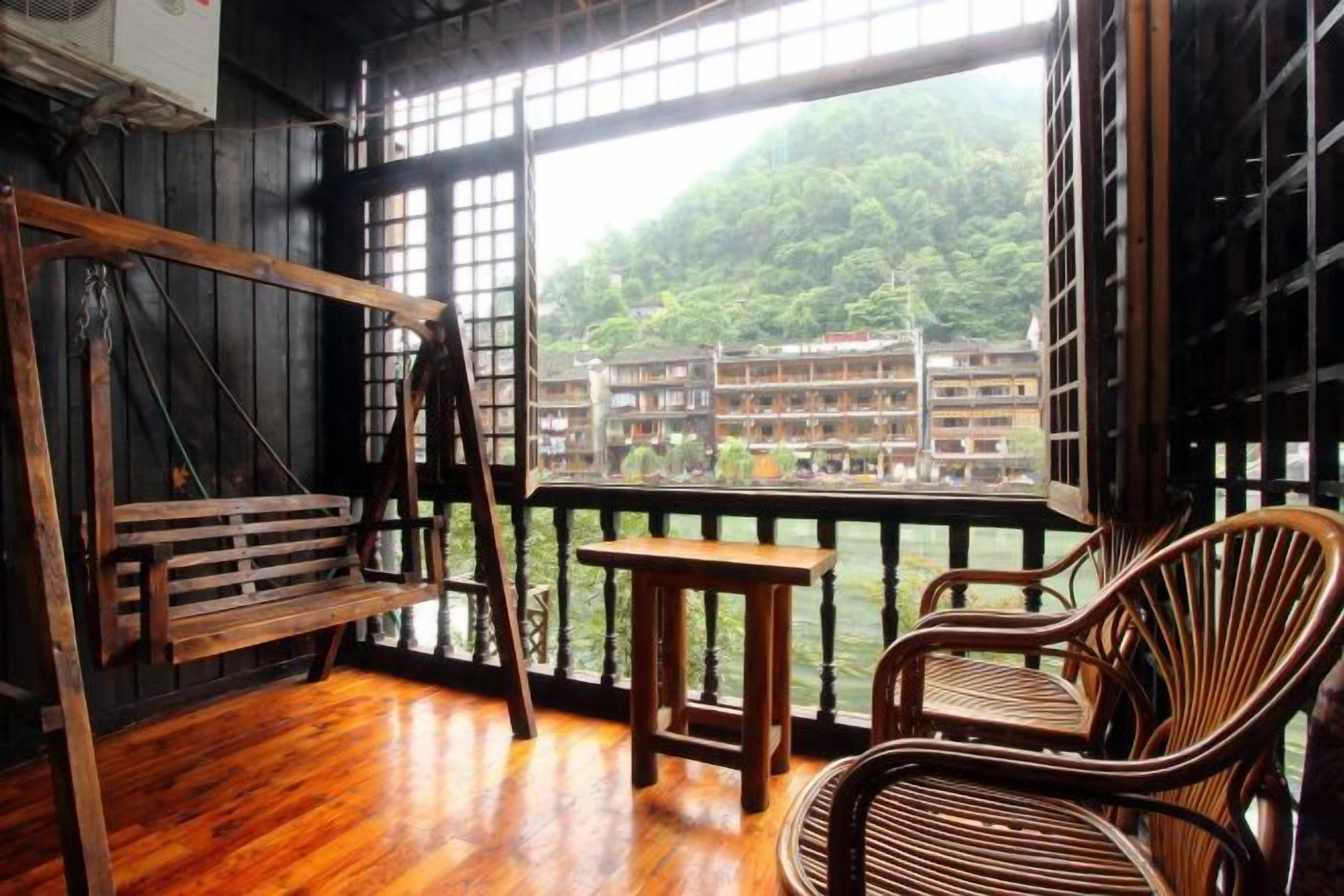 A wooden balcony with a wooden bench and two wicker chairs, overlooking a building and greenery.