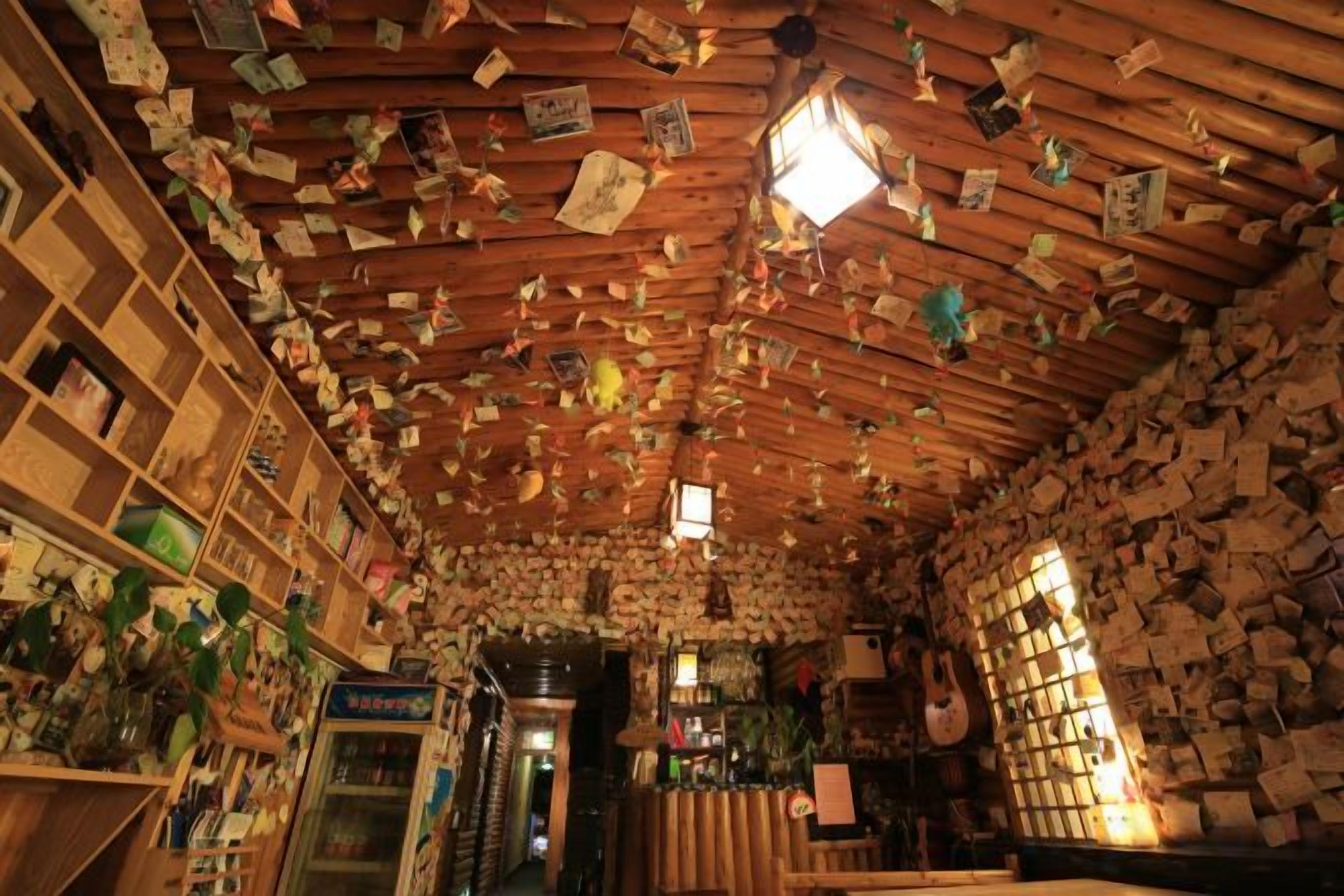 A room with a wooden ceiling covered in numerous papers and notes. There are shelves with various items and a doorway leading to another room.