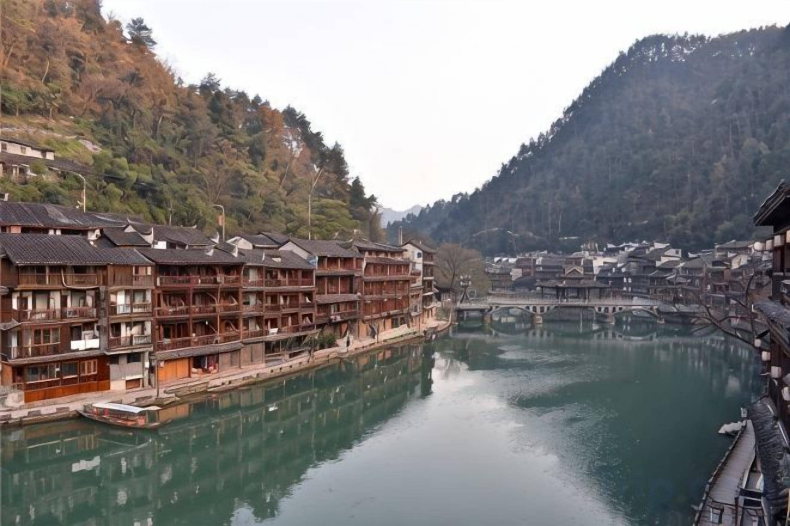 A traditional wooden building with multiple floors, a river in the foreground, and a mountainous backdrop.