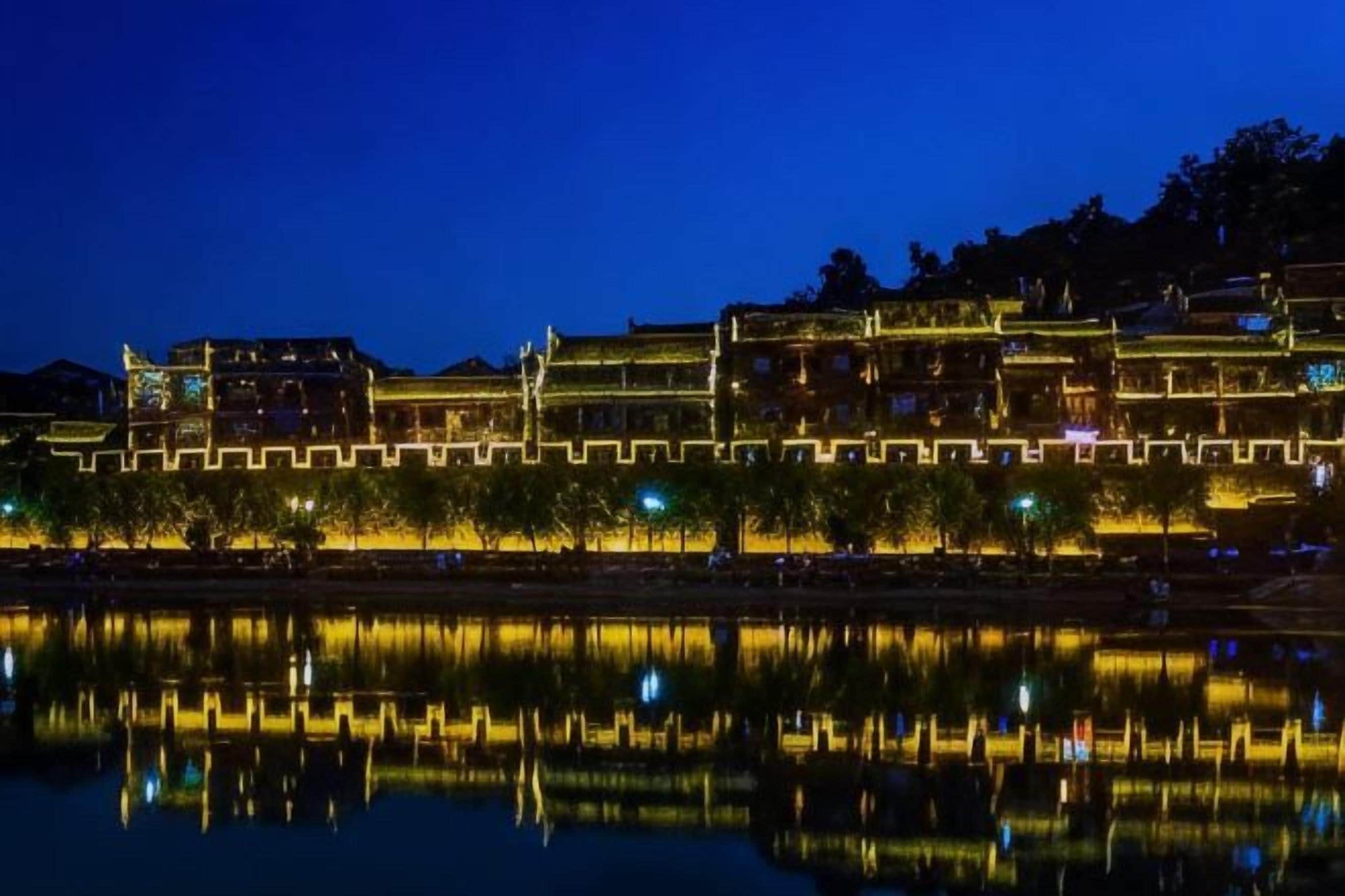 A traditional building with multiple levels, illuminated by warm lighting, reflecting on a calm body of water.