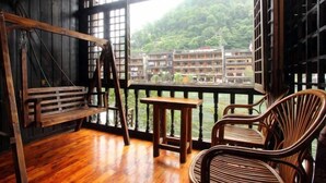 A wooden deck with a bench, a small table, and wicker chairs, overlooking a traditional building with multiple floors and a lush green hillside in the background. - A Good Year (Fenghuang County)