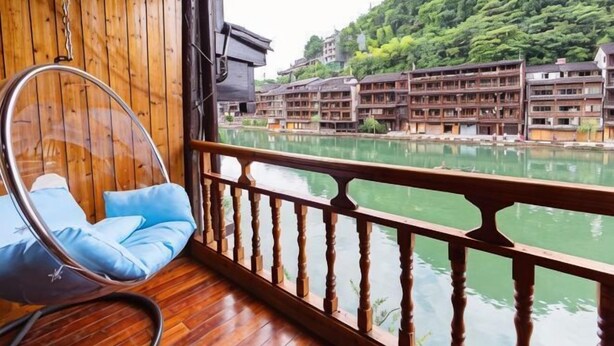 A wooden interior with a hanging chair by a wooden railing overlooking a river and traditional buildings. - A Good Year (Fenghuang County)