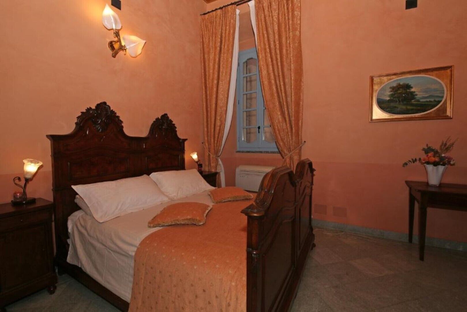A bedroom with a wooden bed, a nightstand with a lamp, a window with curtains, and a painting on the wall.