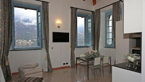 A room with a dining table, chairs, a sofa, and a TV. There are windows with curtains, and a view of a landscape with buildings. - Vice King's Palace (Lezzeno)