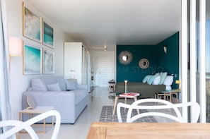 A modern living room with a grey sofa, a dining table with chairs, and a kitchen area visible through an open door.