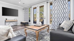A modern living room with a flat-screen TV, a coffee table, a sofa, and a large window with patterned curtains. - Lakefront Gem in Lake Hamilton (Lake Hamilton)
