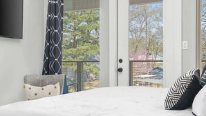 A bedroom with a bed, a chair, a television, and a sliding glass door leading to a balcony with a view of trees and a building. - Lakefront Gem in Lake Hamilton (Lake Hamilton)