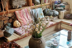 Quarto com parede de pedra, prateleira de madeira com diversos objetos, mesa de vidro com um vaso de flores e um tapete tecido.