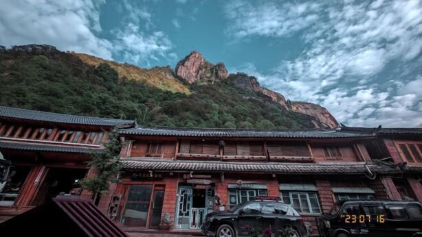 Un edificio tradicional con techo inclinado y estructura de madera, situado en una zona montañosa con una formación rocosa destacada al fondo.