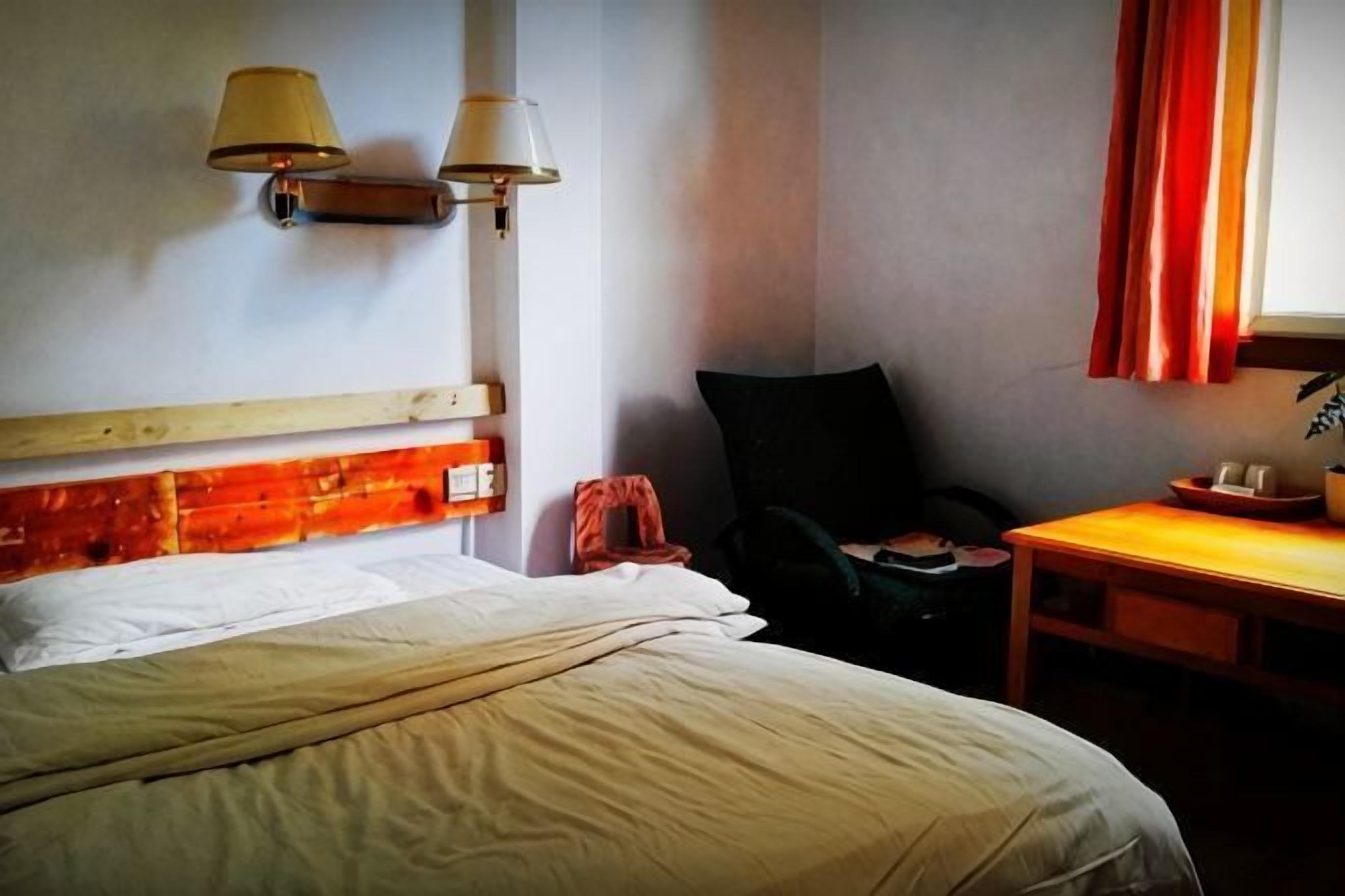 A hotel room with a bed, a chair, a small table, and wall-mounted lamps.