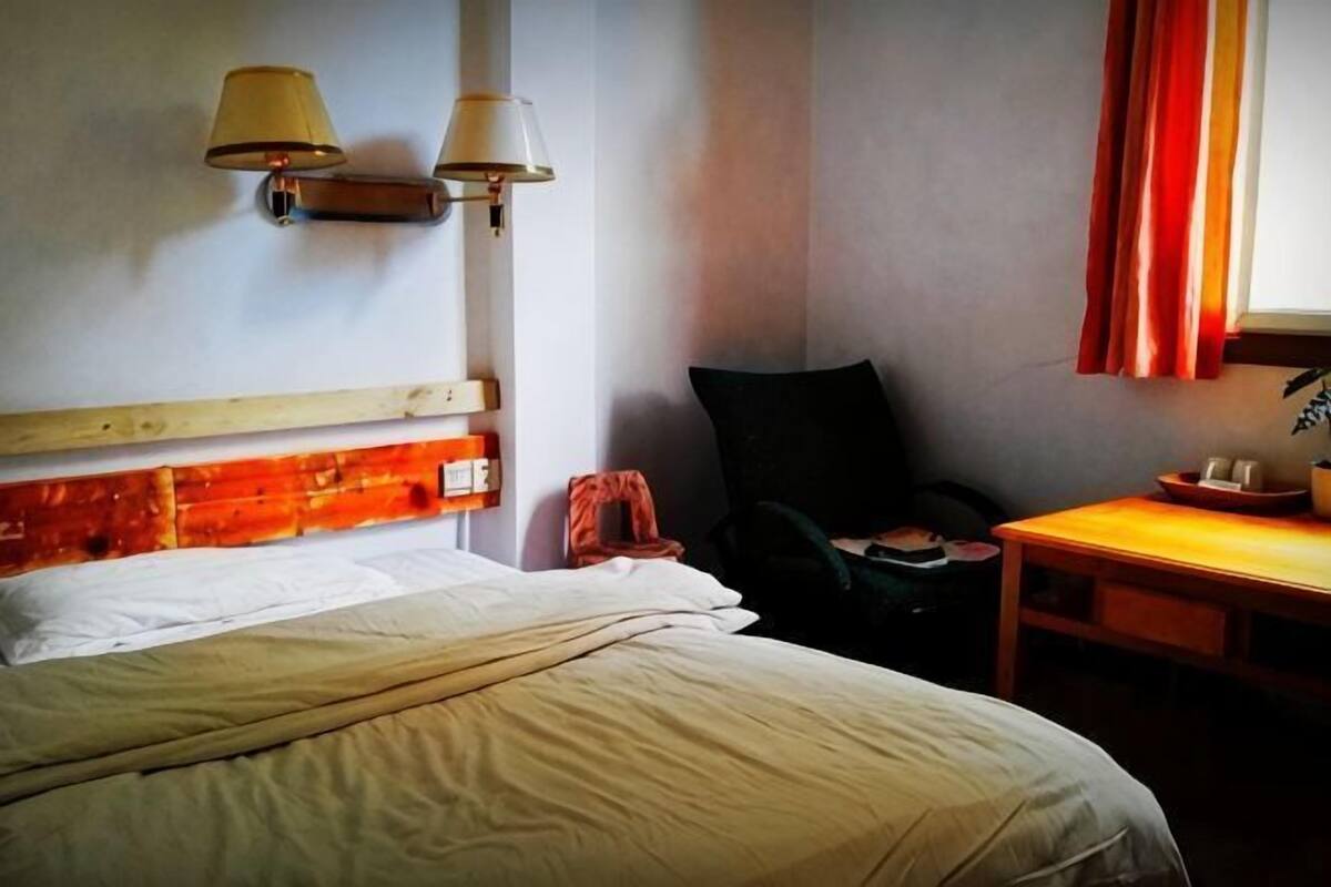 Quarto de hotel com cama, uma cadeira, uma mesinha e luminárias fixadas na parede.