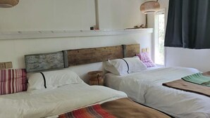 Una habitación con dos camas, cabecero de madera, ropa de cama blanca, una lámpara de mimbre y una ventana con cortina.