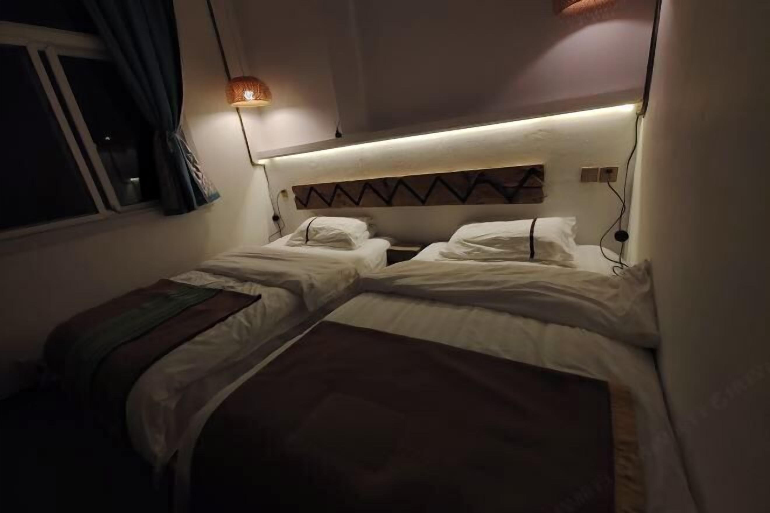 A small, dimly lit room with two single beds, a window with curtains, and a wall-mounted light fixture.
