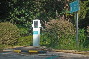 Estación de carga pública para vehículos eléctricos, con pantalla digital, rodeada de vegetación.