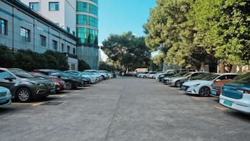Un estacionamiento con varios coches aparcados en filas, un edificio con fachada curva y árboles a lo largo de la calle.