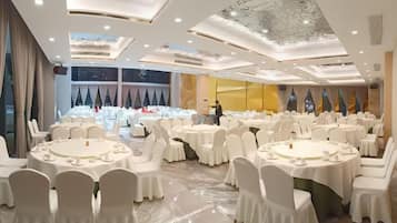 A banquet hall with round tables and white chairs, a high ceiling with intricate designs, and a view of the night sky through large windows.