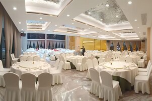 Un salón de banquetes con mesas redondas y sillas blancas, un techo alto con diseños intrincados y vistas del cielo nocturno a través de grandes ventanales.