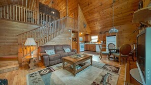 A spacious log cabin interior with a living room, kitchen, and a staircase leading to an upper level.