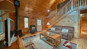 Interior acogedor de una cabaña de troncos con una escalera de madera, un sofá, un televisor y una chimenea.