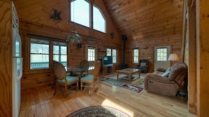A cozy cabin interior with wooden walls, a dining area with chairs and a table, a sofa, a fireplace, and a television.