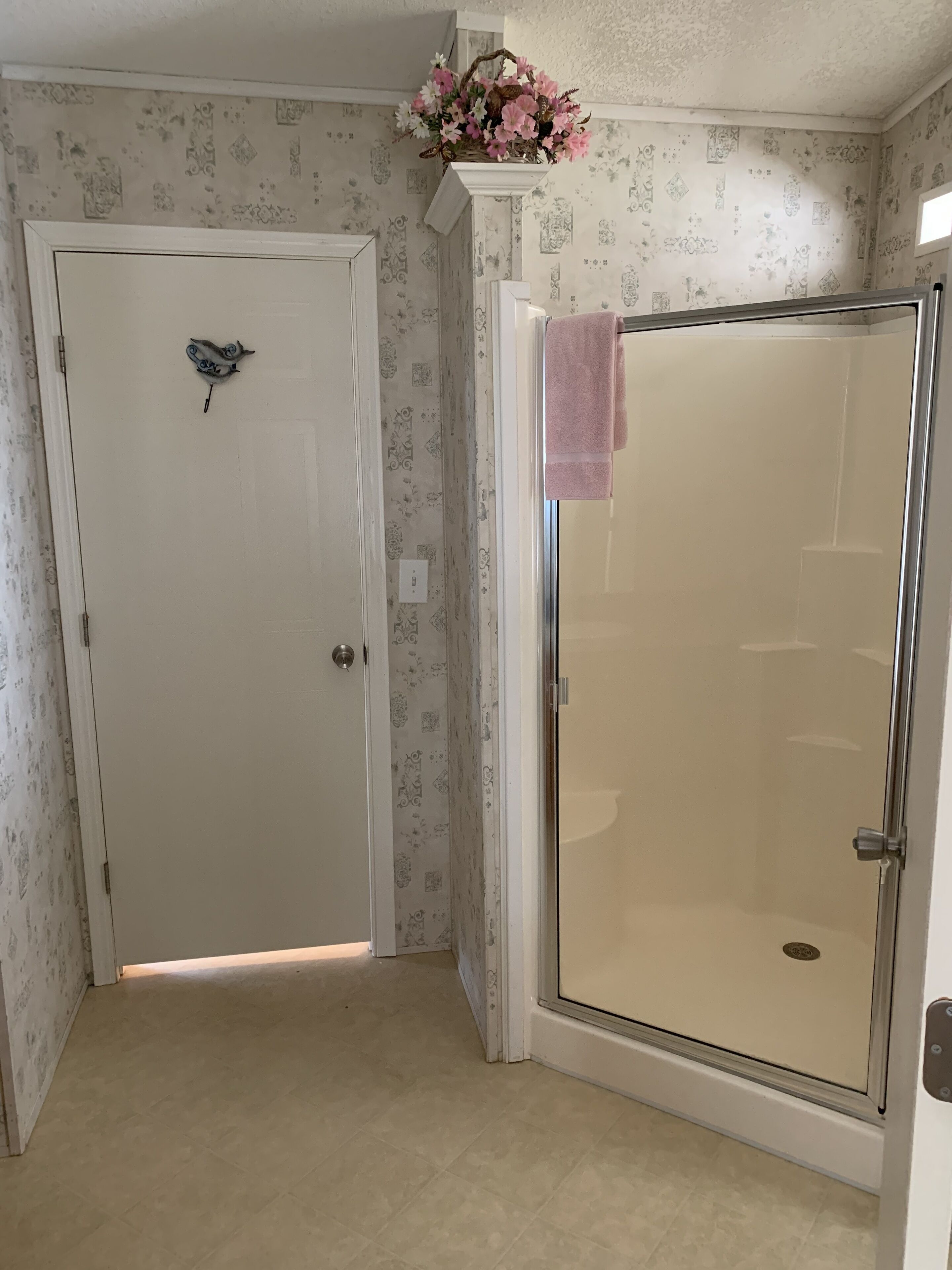 A room with a white door, a shower with a pink towel, and patterned wallpaper.