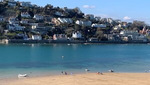 A coastal beach with clear water, a sandy shore, and buildings in the background.