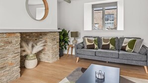 A living room with a stone fireplace, a grey sofa with patterned cushions, a round mirror, and a window with a view of a brick building.