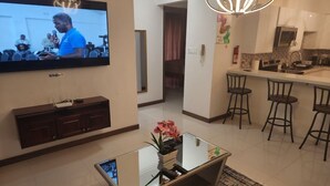 Uma sala de estar moderna com televisão de tela plana, uma mesa de centro de vidro com um vaso de flores, uma área de cozinha com banquetas e uma mesa de jantar.