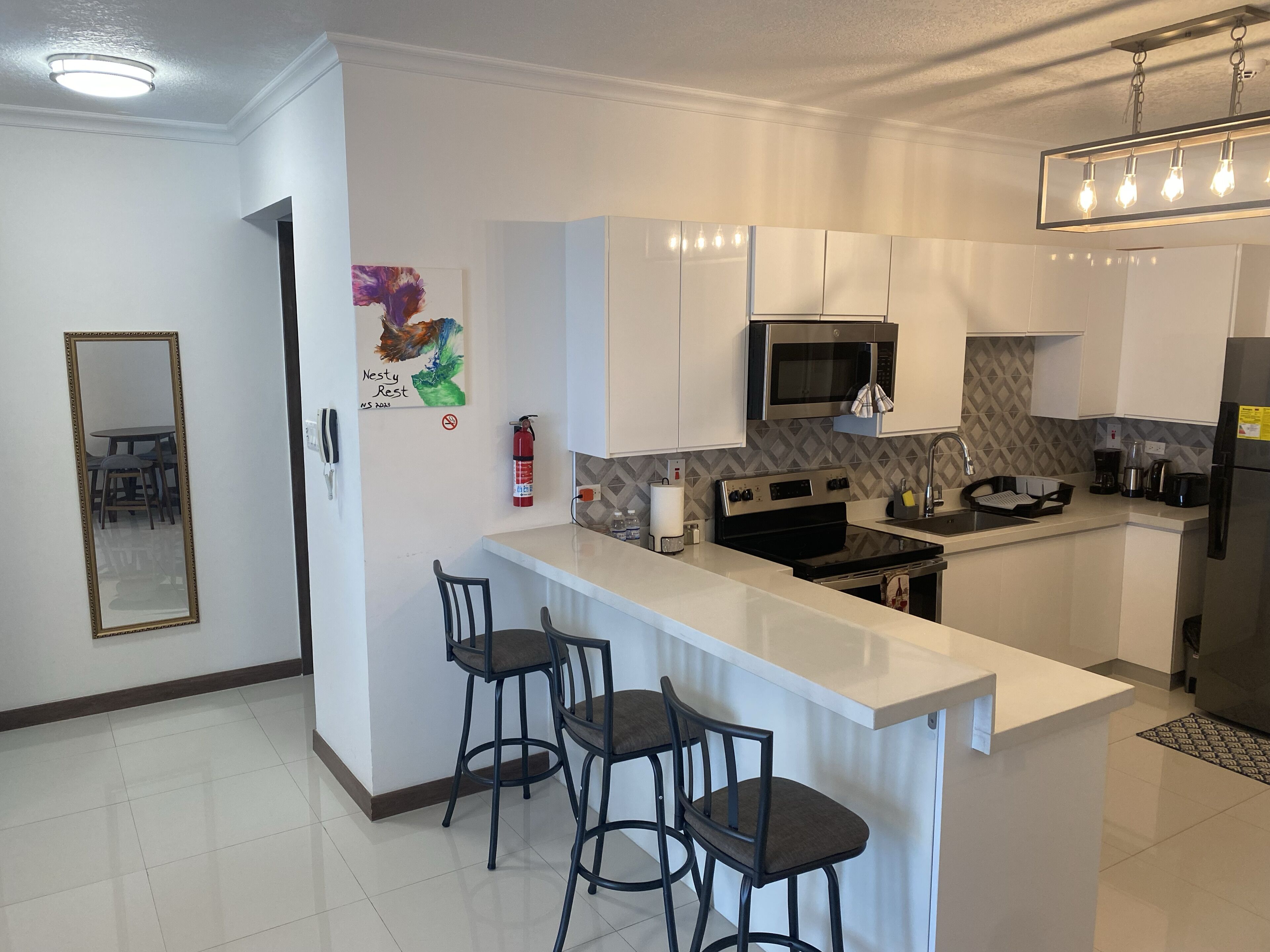 Een moderne keuken met witte kasten, een roestvrijstalen magnetron, een kookplaat en een koelkast. Er staan twee barkrukken en zit een brandblusser aan de muur.