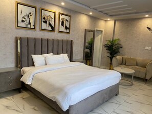 A modern bedroom with a large bed, a grey upholstered sofa, a small round table, and three abstract paintings on the wall. - Luxus Suites (Lahore)