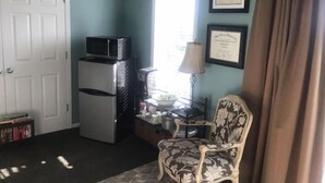 Quarto compacto com porta branca, refrigerador branco, micro-ondas, poltrona estampada, abajur e dois certificados emoldurados na parede.