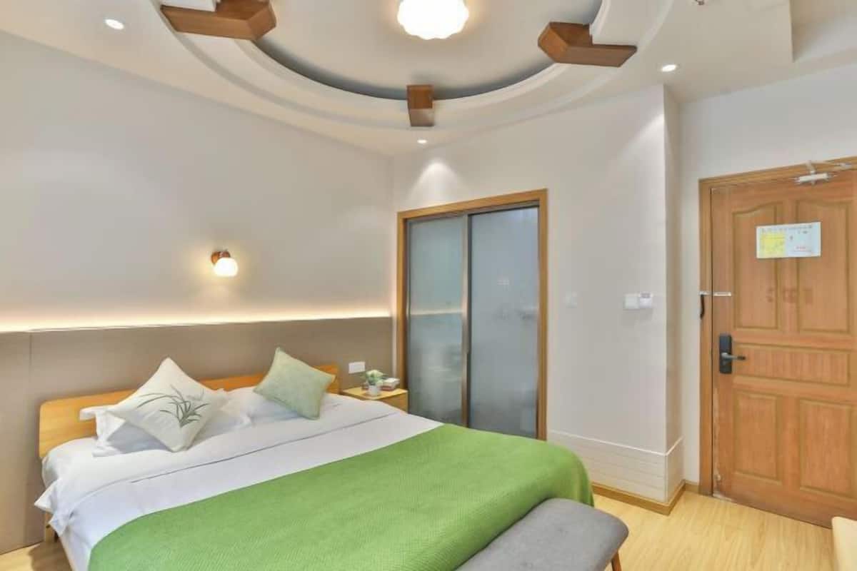 A hotel room with a bed, a nightstand, a green throw blanket, a wooden door, and a ceiling light fixture.