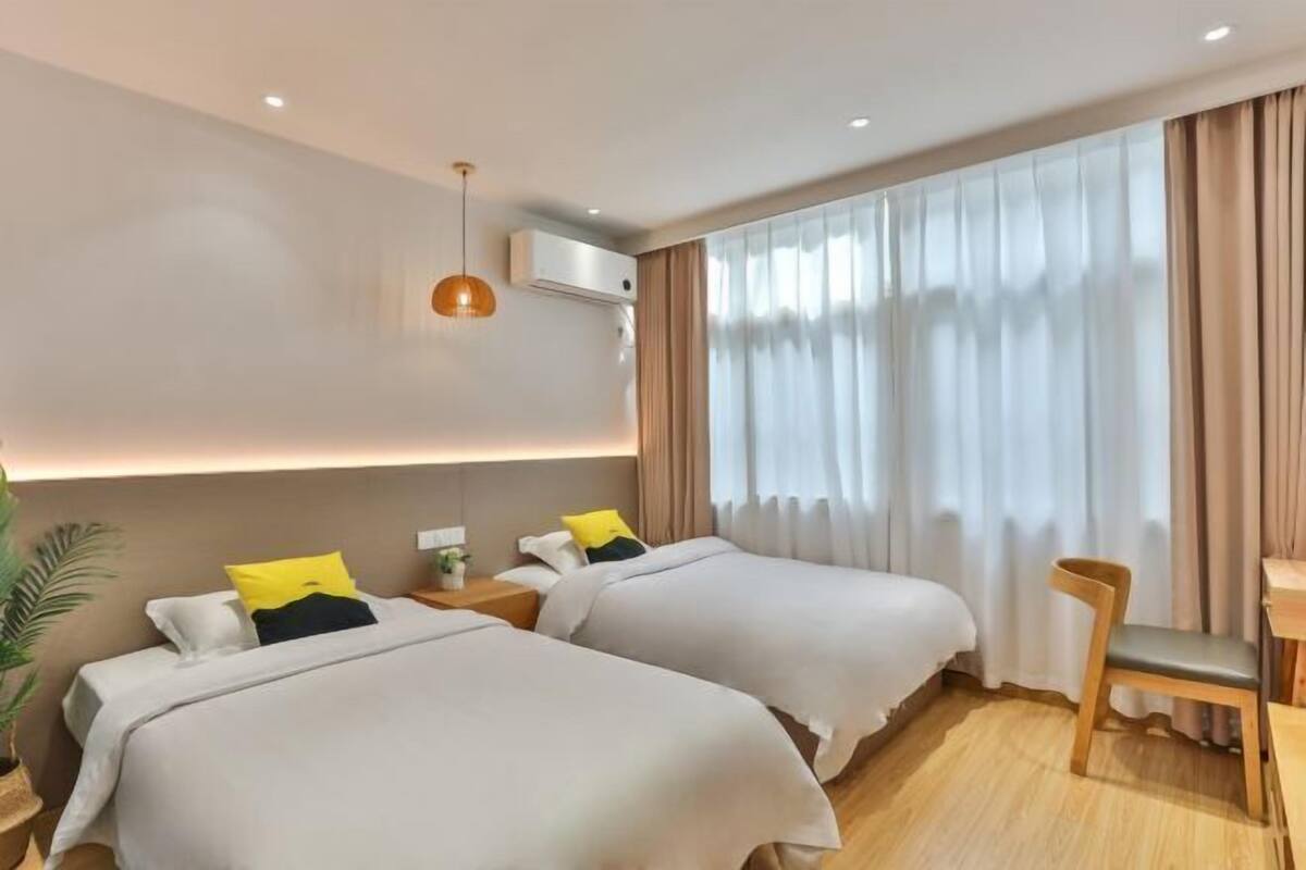 A hotel room with two beds, a desk, a chair, a potted plant, and a large window with curtains.