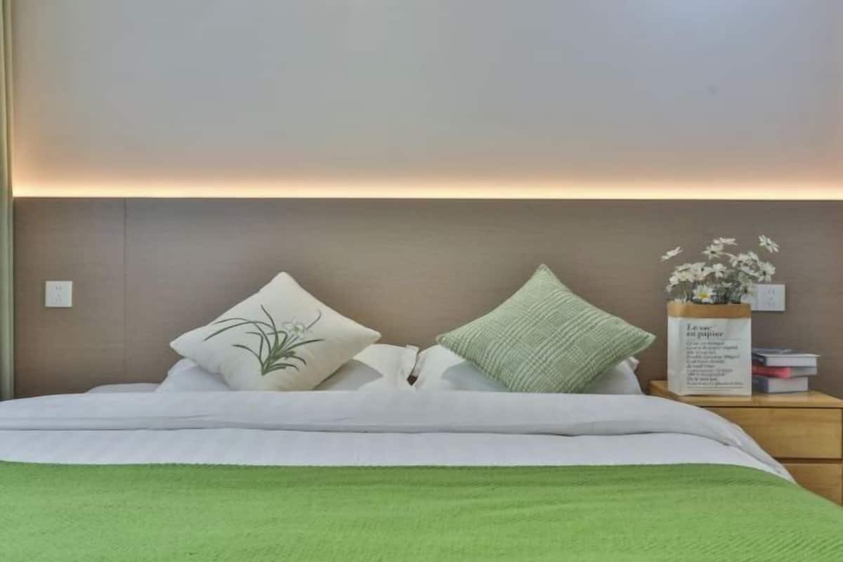 A neatly made bed with white and green bedding, a bedside table with a vase of flowers, and a wall-mounted light fixture.