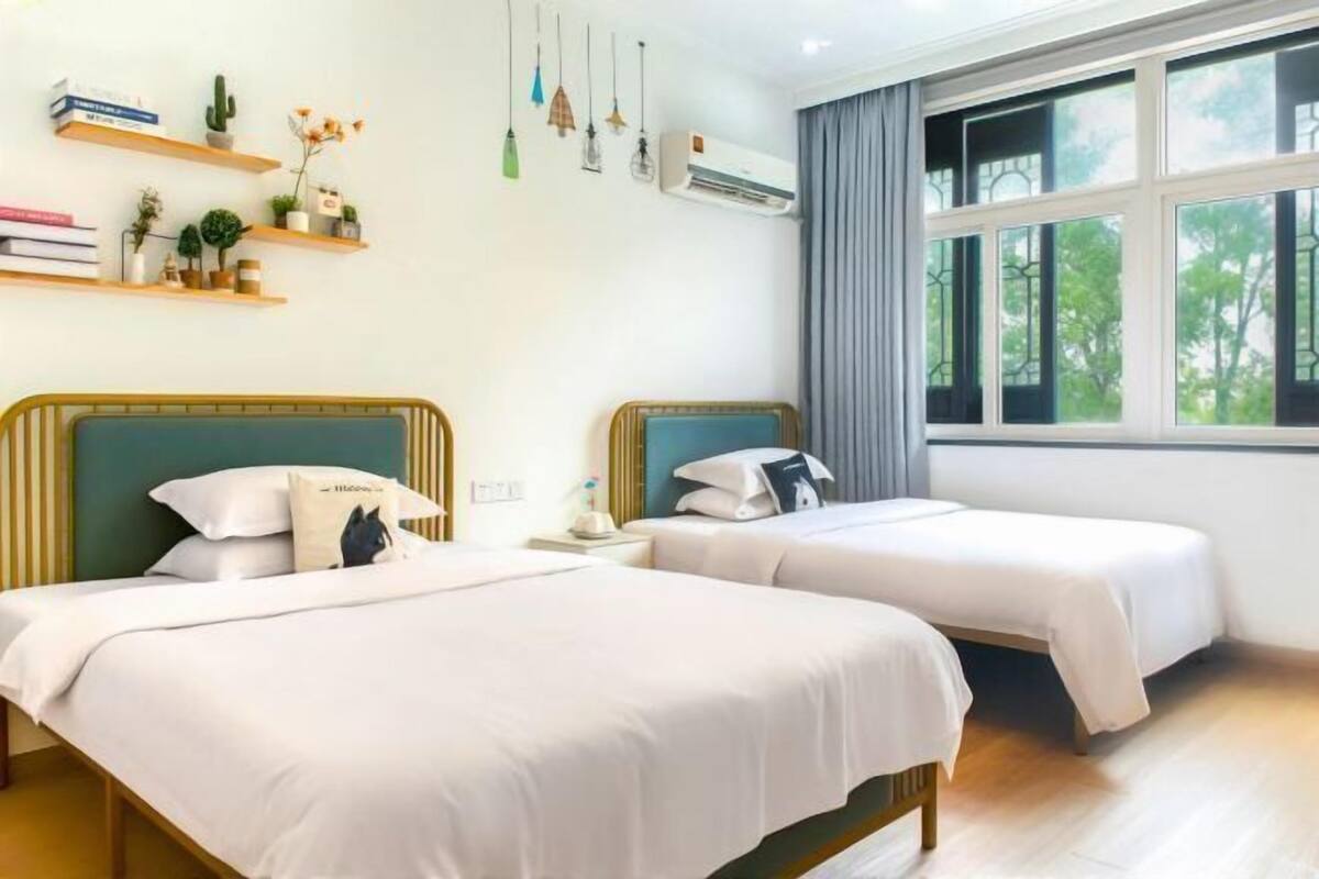 A hotel room with two beds, a window with curtains, a wall-mounted air conditioner, and shelves with books and plants.
