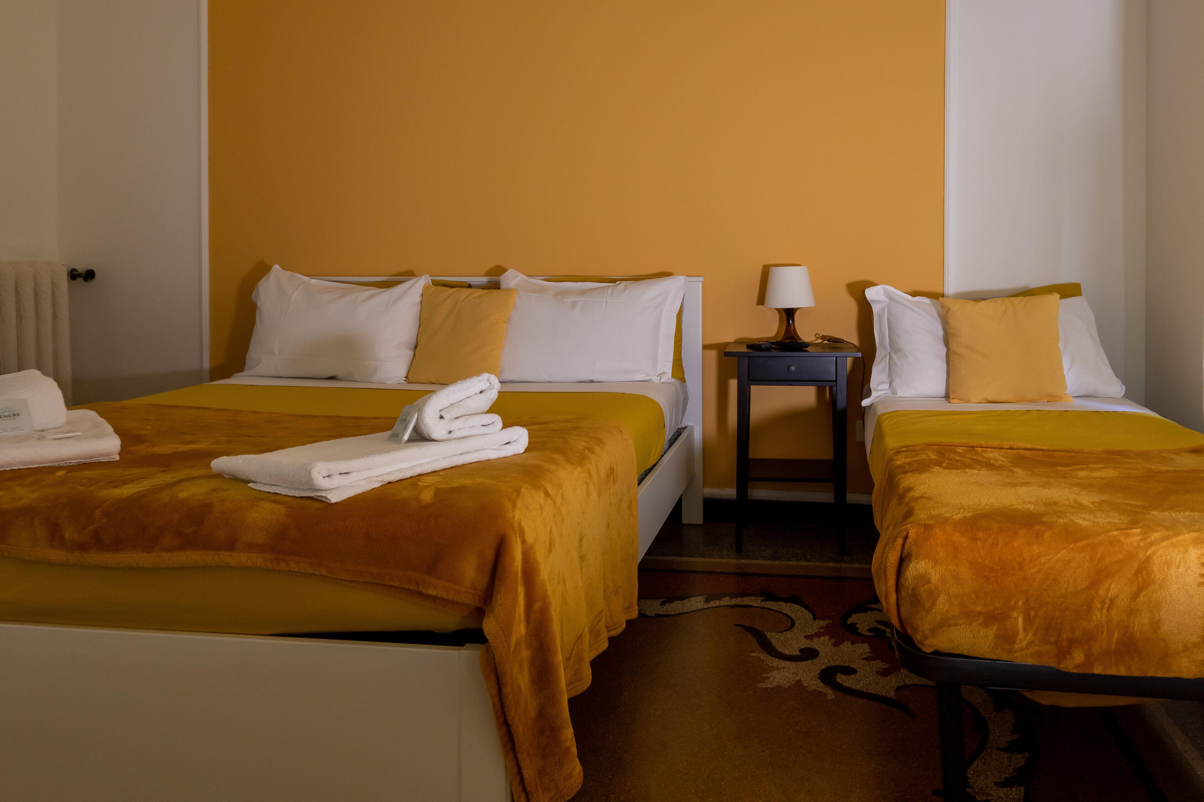 Een hotelkamer met twee bedden, elk met een gele sprei en witte kussens. Daartussen staat een nachtkastje met een lamp.
