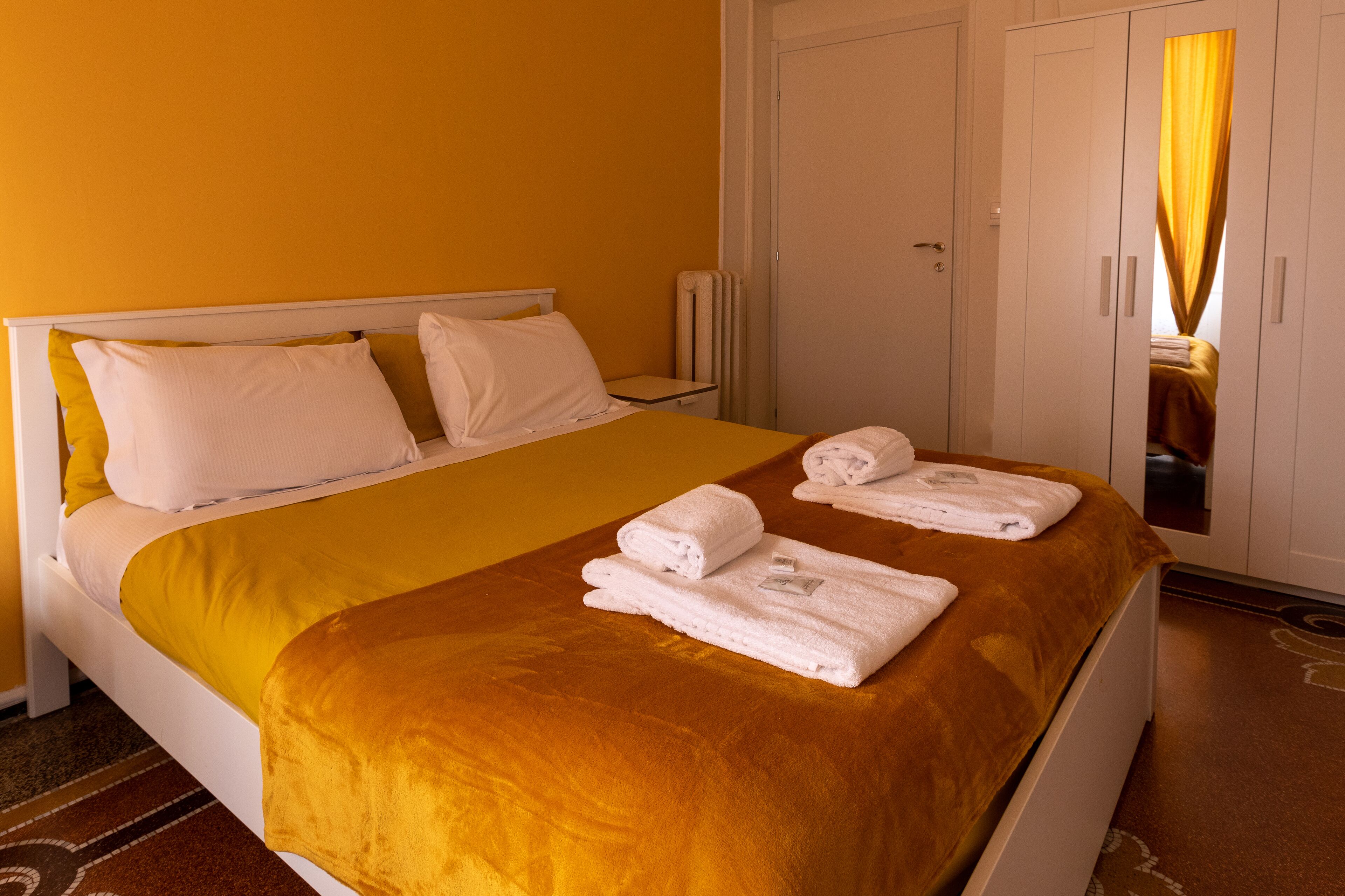 A hotel room with a bed, white headboard, yellow walls, and a wardrobe with a mirror.