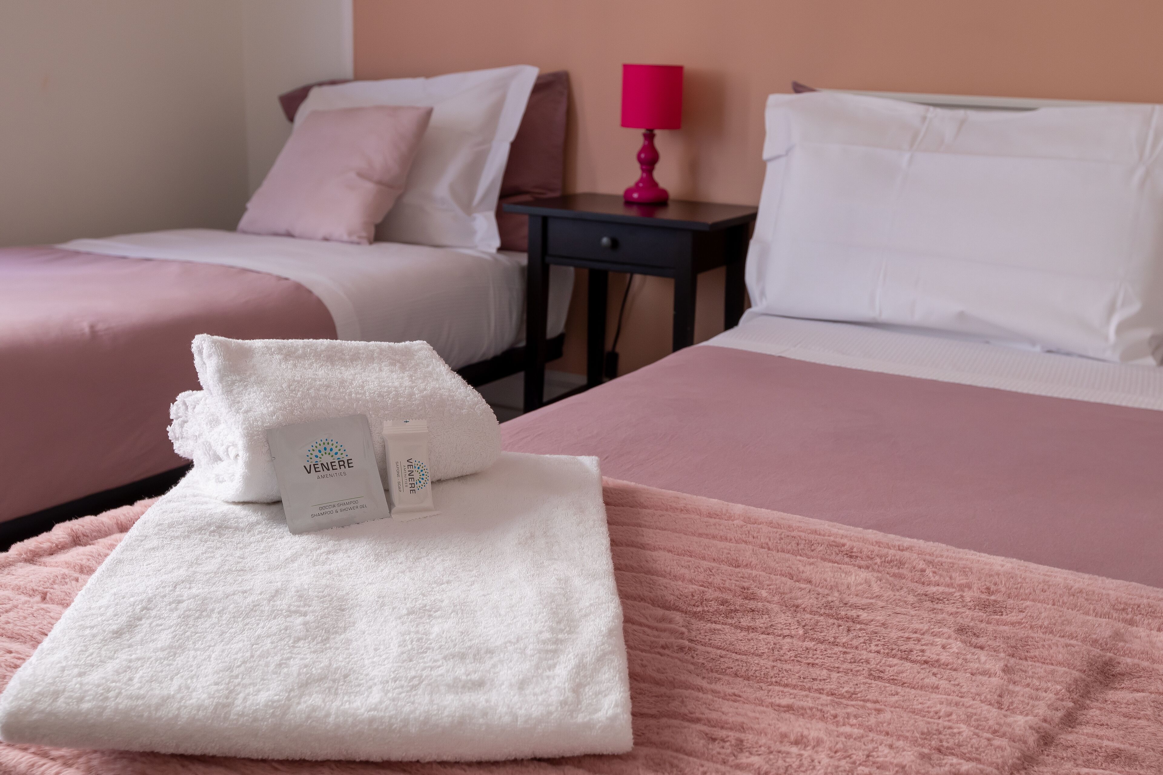 Ein Hotelzimmer mit zwei Betten, eines mit einer rosa Decke und das andere mit einer weißen. Auf dem Bett mit der rosafarbenen Decke liegt ein weißes Handtuch mit einem Vénere-Etikett. Ein rosa Lampenschirm steht auf einem Nachttisch neben dem Bett mit der weißen Decke.