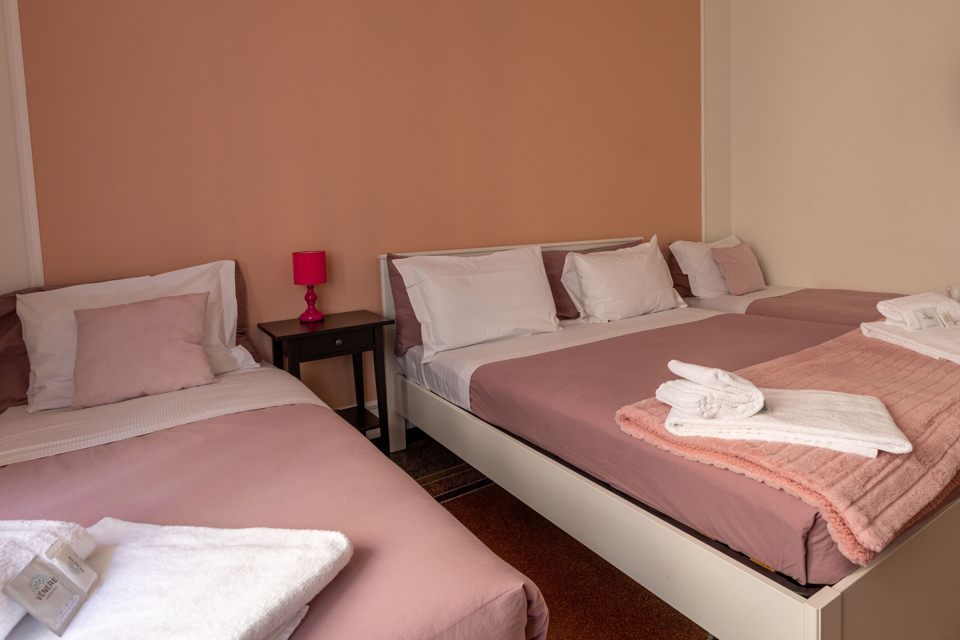 A hotel room with two beds, each with pink bedding and white pillows. There is a small table with a pink lamp and a bottle of lotion.