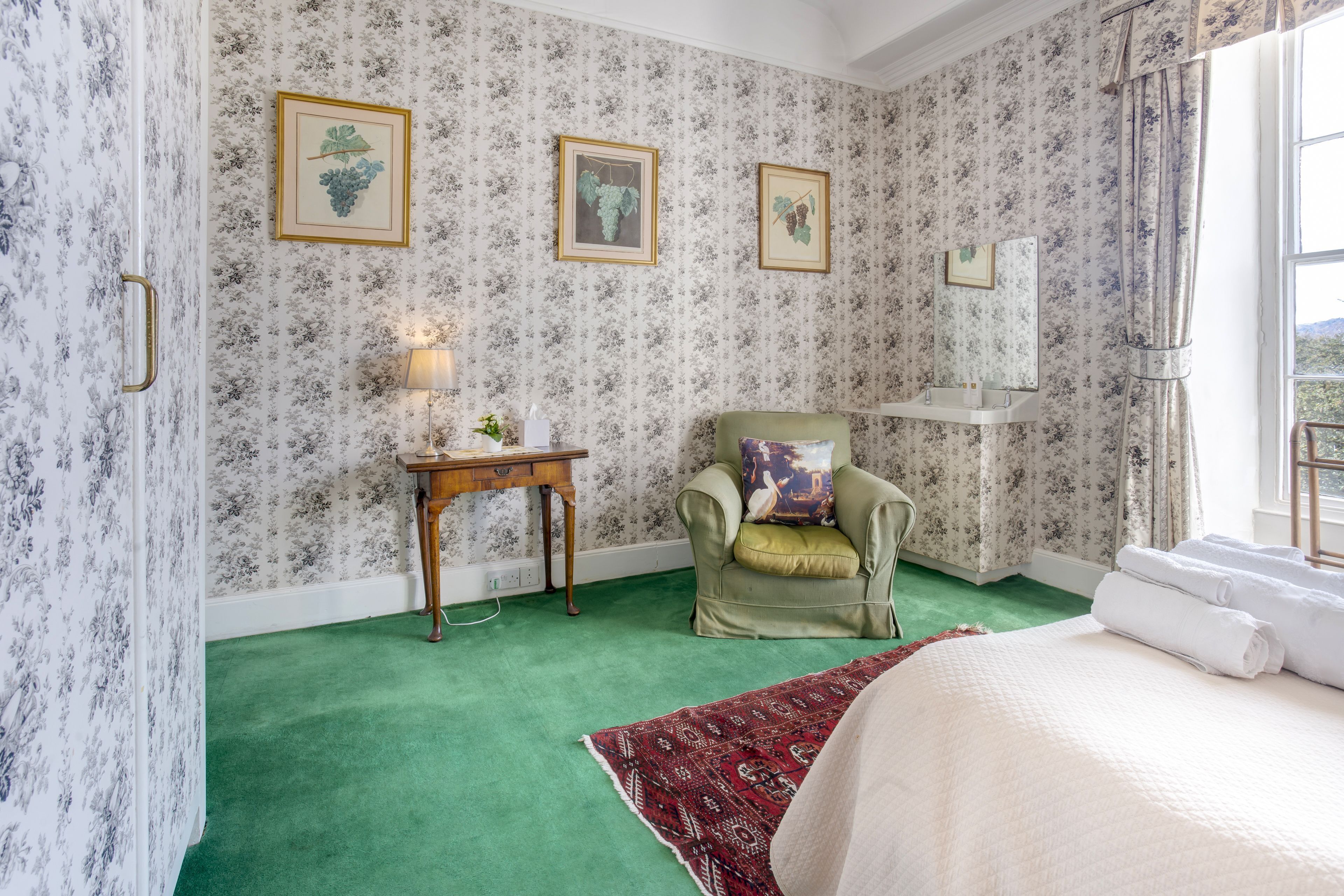 A room with floral wallpaper, a green carpet, a green armchair with a cushion, a small wooden table with a lamp, and a bed with white linens.