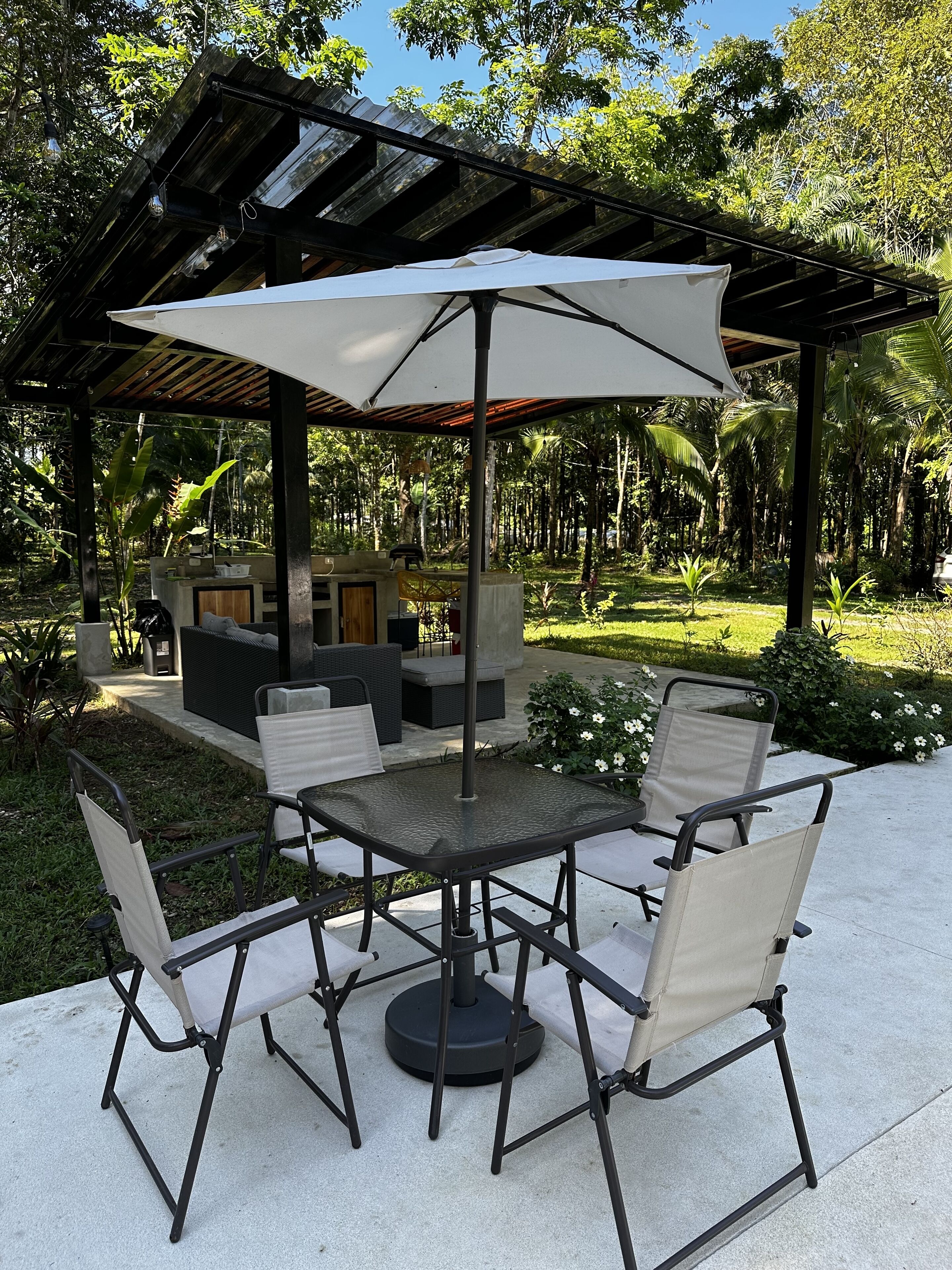 An outdoor patio with a round table and six chairs, surrounded by a pergola and lush greenery.