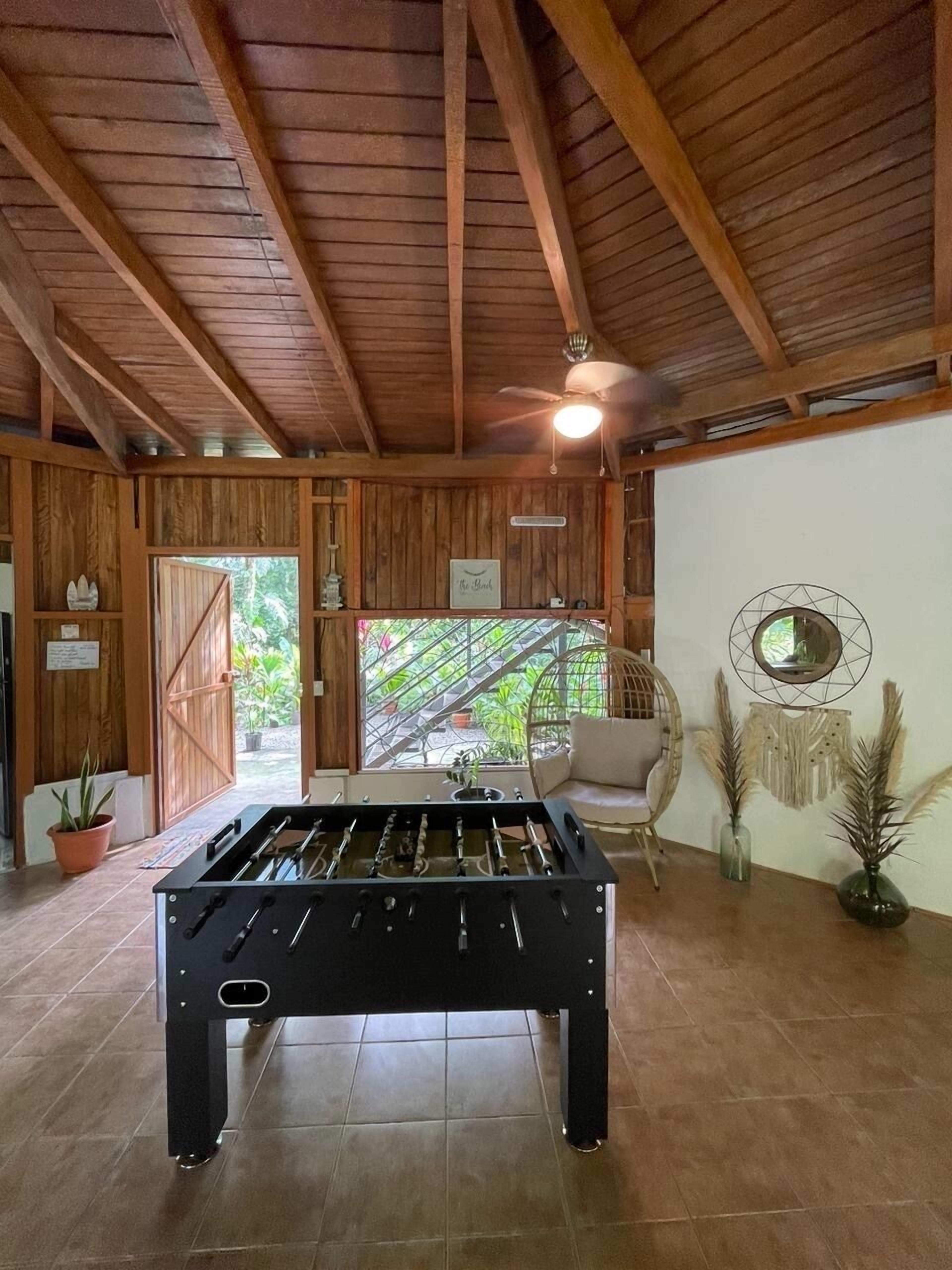 Una stanza con tavolo da calcio, travi in legno a soffitto, ventilatore a soffitto e vista attraverso una porta vetrata su una zona esterna con piante.