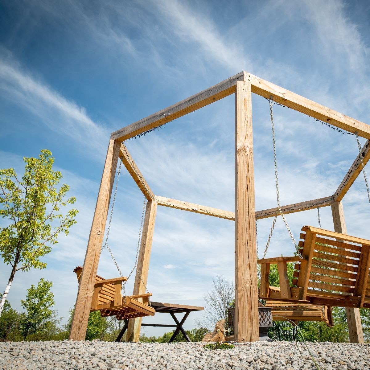 A wooden swing set with a table and chairs, surrounded by gravel and trees.