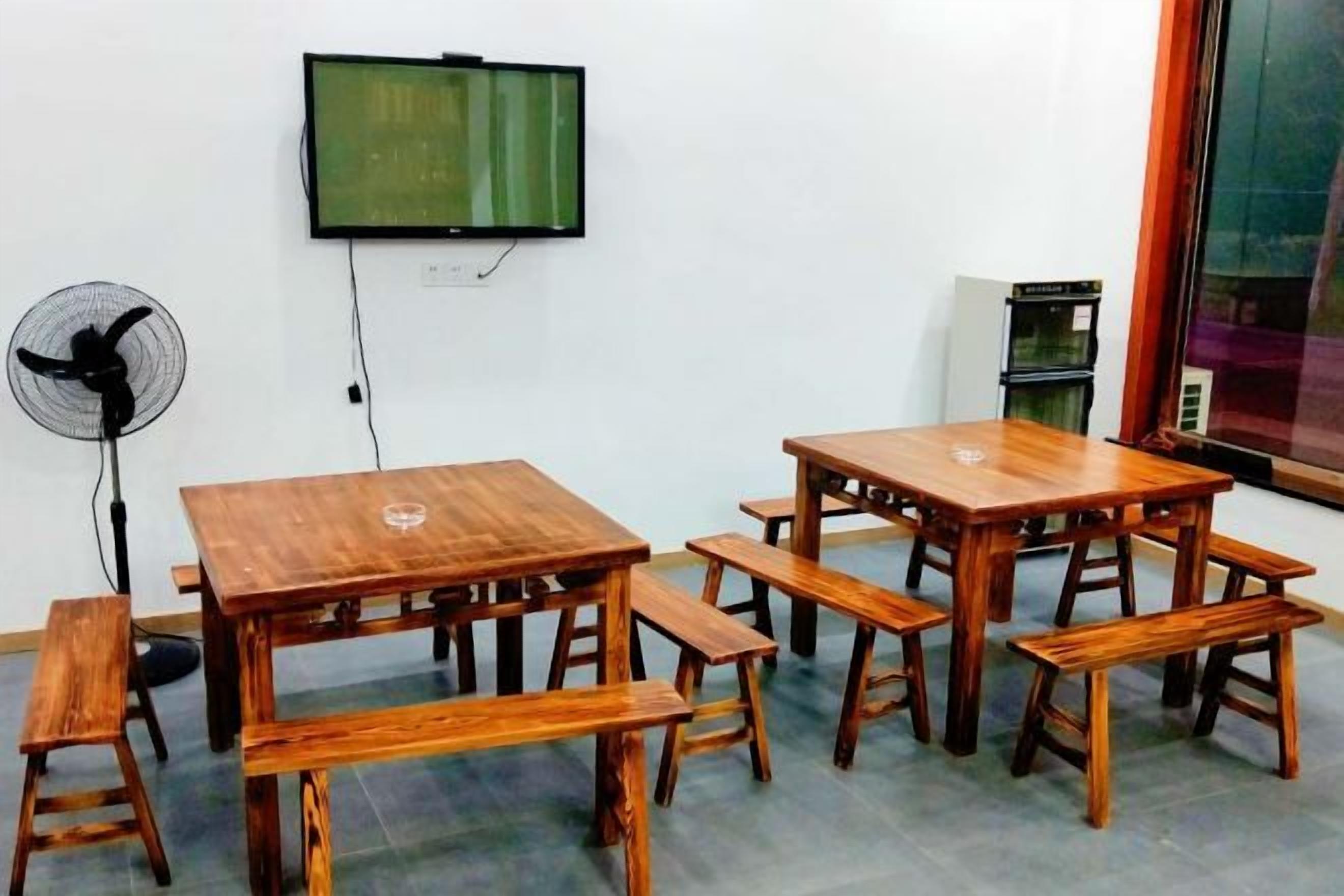 Una habitación con una mesa de madera y bancos, un televisor montado en la pared, un ventilador y una ventana.