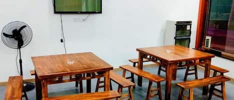 A room with a wooden table and benches, a wall-mounted TV, a fan, and a window.