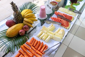 Una mesa de buffet con diversas frutas y verduras, entre ellas plátanos, mangos y zanahorias.