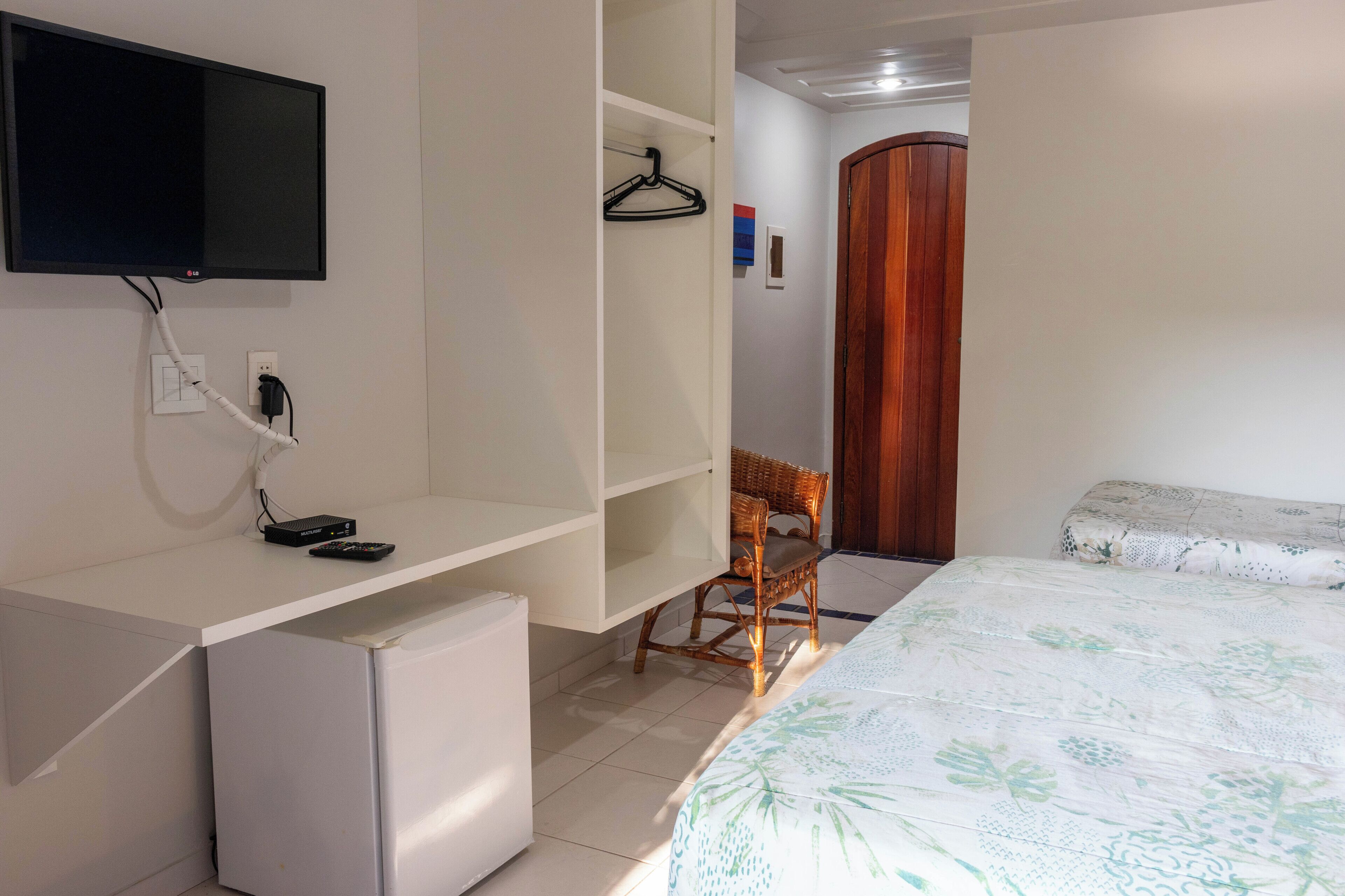 A bedroom with a bed, a desk, a small refrigerator, a television mounted on the wall, and a wooden door.
